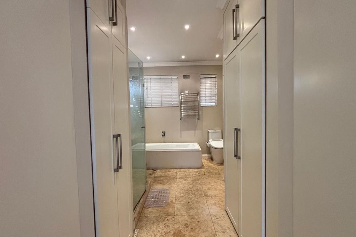 A modern bathroom featuring built-in storage cupboards, a glass-enclosed shower, a bathtub, and a heated towel rail with neutral tiled flooring.