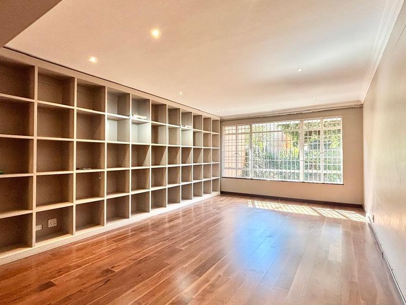 Spacious living area featuring polished hardwood flooring, extensive built-in shelving, and large windows with security bars, offering a bright and versatile space.