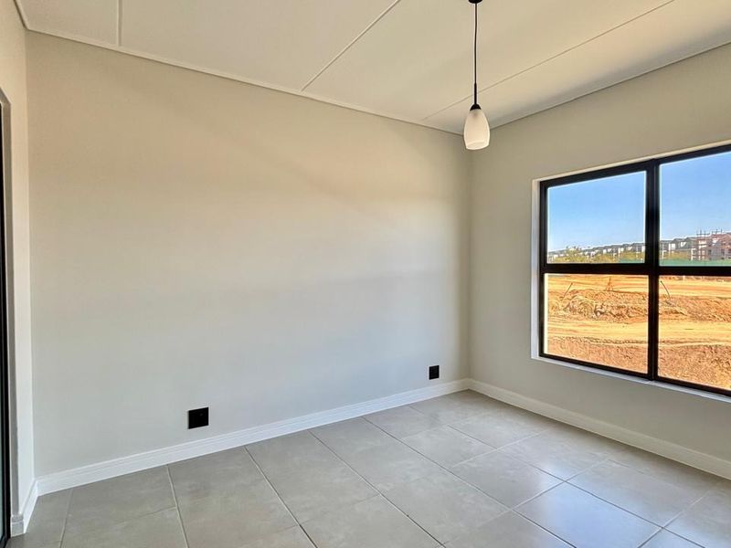 A bright, modern, and empty bedroom featuring neutral tiled flooring, clean finishes, and large black-framed windows with an exterior view.