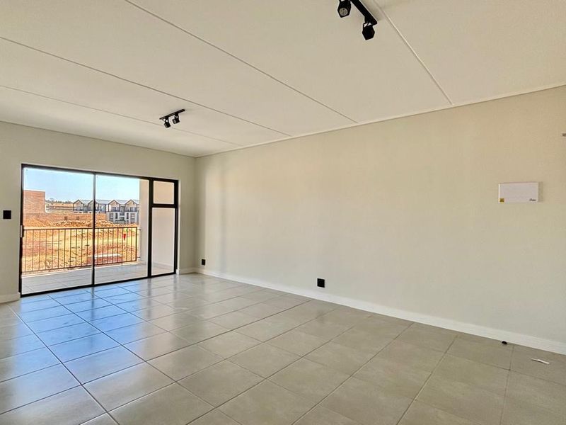 A bright, open-plan living area featuring neutral tiled flooring, modern track lighting, and a large sliding glass door leading to an outdoor balcony.