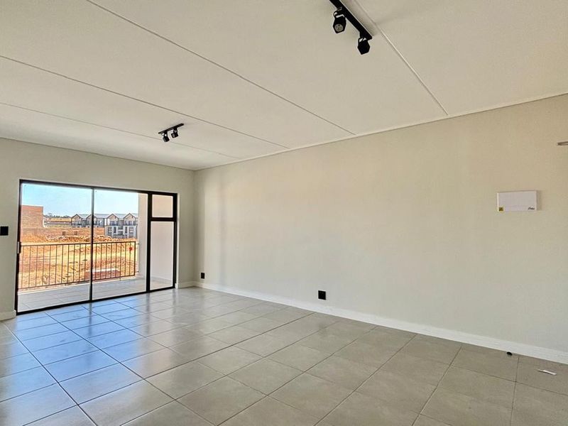 Spacious, empty living area featuring neutral tiled flooring, modern track lighting, and a large sliding glass door providing access to a private balcony.