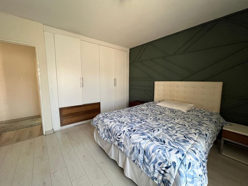 A well-maintained bedroom featuring light laminate flooring, a large built-in wardrobe with integrated drawers, and a modern dark green geometric feature wall.