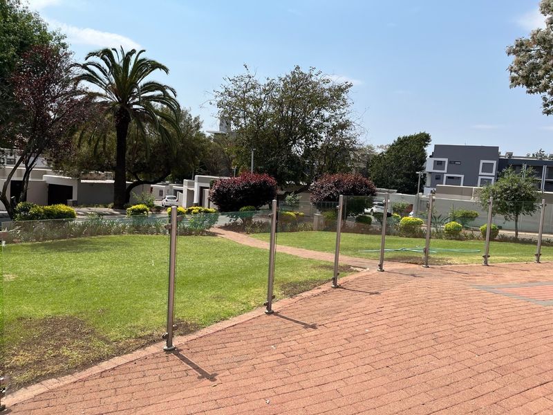 A well-maintained outdoor patio area featuring brick paving and a modern glass balustrade overlooking a landscaped garden and residential complex.