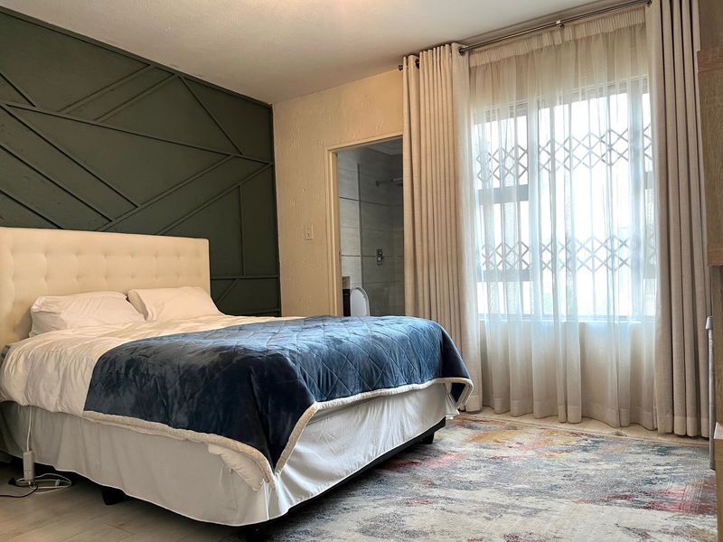 A well-maintained bedroom featuring decorative wall paneling, tiled flooring, and an en-suite bathroom access, with natural light provided by a large window.