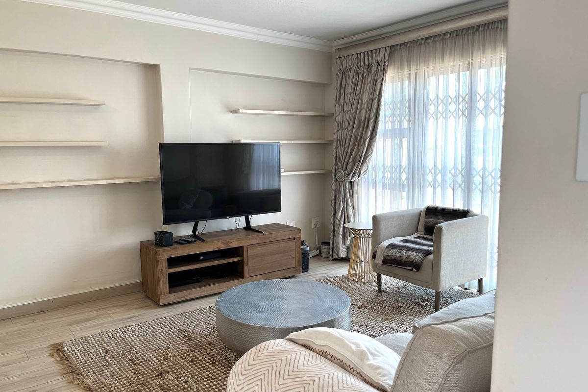A comfortable living room featuring built-in wall shelving, laminate flooring, and large windows with security gates, offering a bright and functional space.