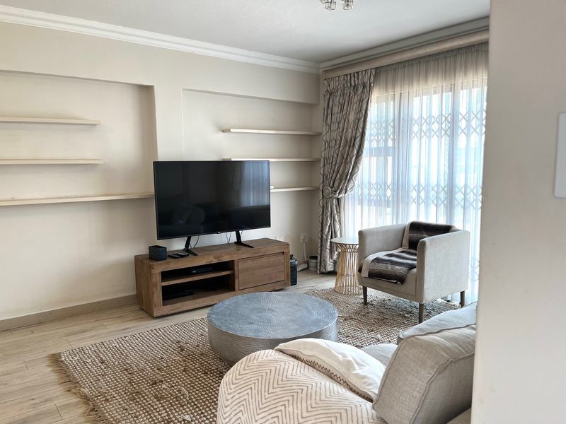 A comfortable living room featuring built-in wall shelving, laminate flooring, and large windows with security gates, offering a bright and functional space.