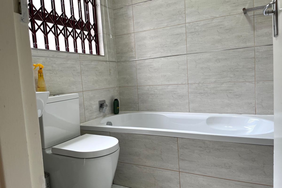 A clean, modern bathroom featuring neutral wall tiles, a built-in bathtub, and a standard toilet, complemented by a window with a security gate.