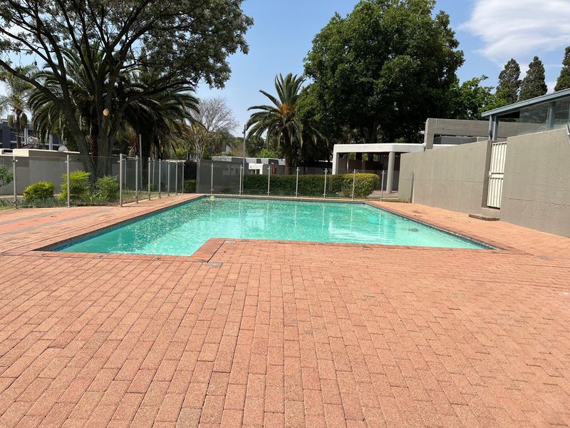 A well-maintained outdoor swimming pool area featuring brick paving, glass safety fencing, and established garden surroundings within the townhouse complex.