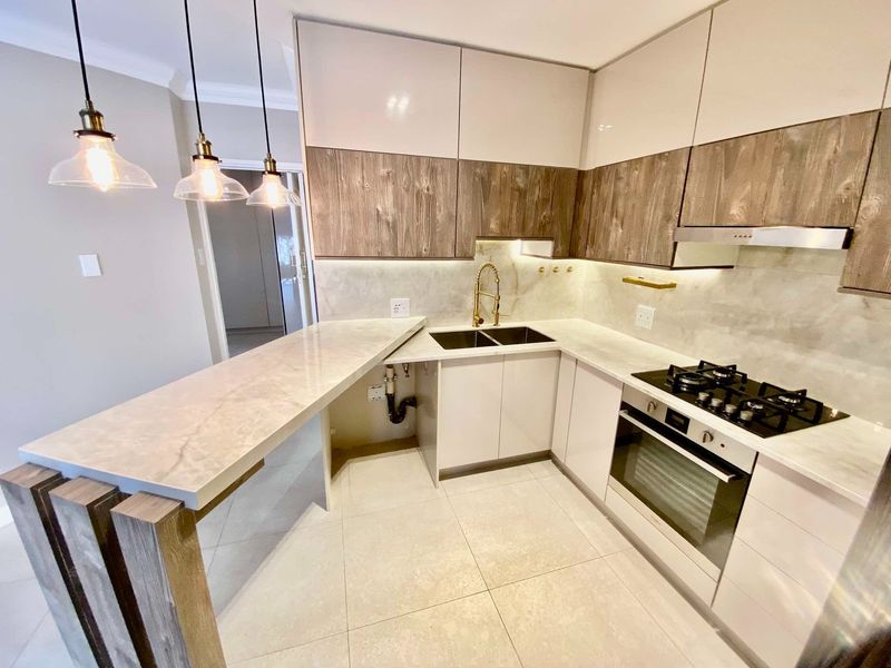 Modern kitchen featuring two-tone cabinetry, stone countertops, a breakfast bar, integrated gas hob, oven, and stylish brass fixtures with under-cabinet lighting.