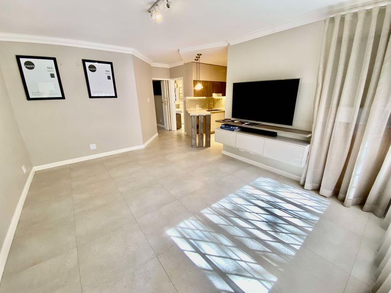 A bright open-plan living area featuring large format floor tiles, coving, and a seamless transition to a modern kitchen with a breakfast bar.
