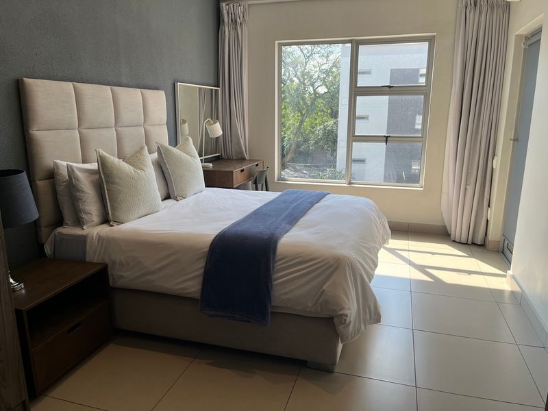 A well-lit bedroom featuring large windows, neutral tiled flooring, and a modern grey accent wall.