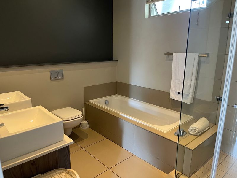 Modern bathroom featuring a double vanity, built-in bathtub with tiled surround, wall-mounted toilet, and a glass shower screen.