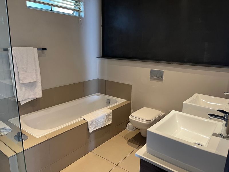Modern bathroom featuring a built-in bathtub, wall-mounted toilet, and dual vessel sinks with contemporary chrome fixtures and neutral tiling.
