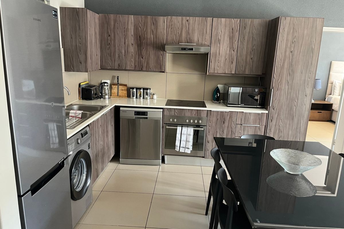 Modern kitchen featuring wood-grain cabinetry, stainless steel appliances, including a built-in oven, hob, extractor fan, dishwasher, and washing machine, with tiled flooring.