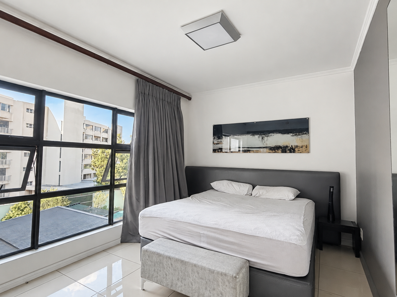 A bright and modern bedroom featuring large windows with natural light, tiled flooring, and a built-in mirrored wardrobe.