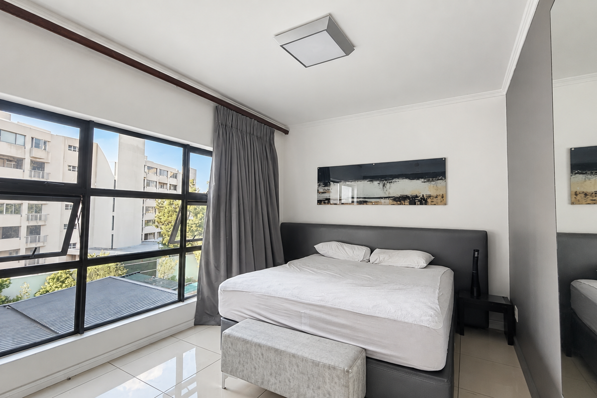 A bright and modern bedroom featuring large windows with natural light, tiled flooring, and a built-in mirrored wardrobe.