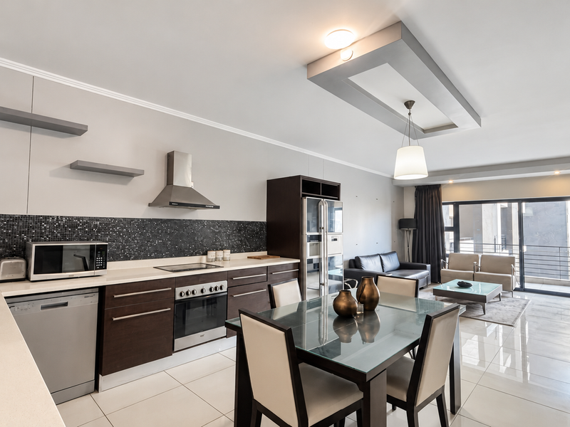 Modern open-plan living area featuring a sleek kitchen with dark cabinetry, mosaic backsplash, integrated appliances, and seamless flow to a bright balcony.
