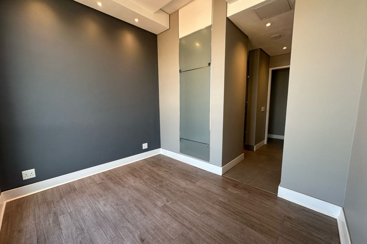 A modern, empty bedroom featuring dark accent walls, recessed lighting, air conditioning, and high-quality laminate flooring, leading into a tiled hallway area.