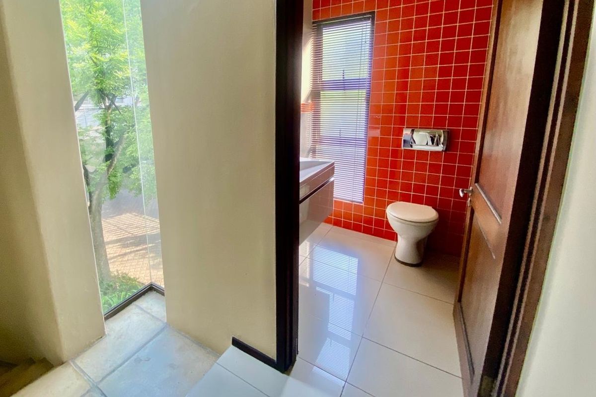 A bright bathroom featuring a wall-mounted toilet with a flush plate, tiled flooring, and a vanity basin, complemented by a large window with blinds.