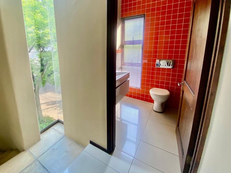 A bright bathroom featuring a wall-mounted toilet with a flush plate, tiled flooring, and a vanity basin, complemented by a large window with blinds.