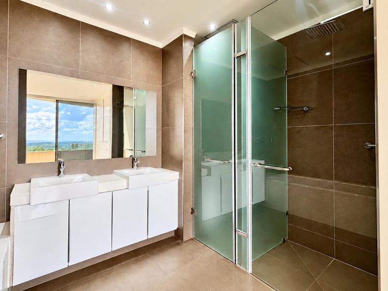 Modern bathroom featuring a double vanity, large wall mirror, and a spacious glass-enclosed shower with a rain shower head and contemporary brown tiling.
