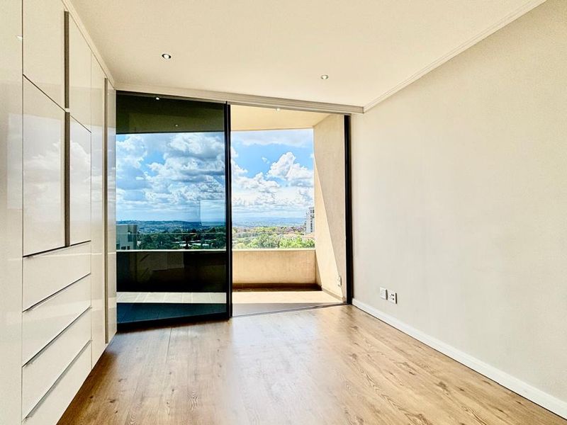 A bright, modern bedroom featuring sleek built-in cupboards, laminate flooring, and a large sliding glass door opening onto a private balcony with scenic views.