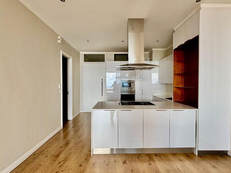 Modern kitchen featuring a central island, built-in oven, extractor fan, and sleek white cabinetry with integrated shelving, set against light wood-look flooring.