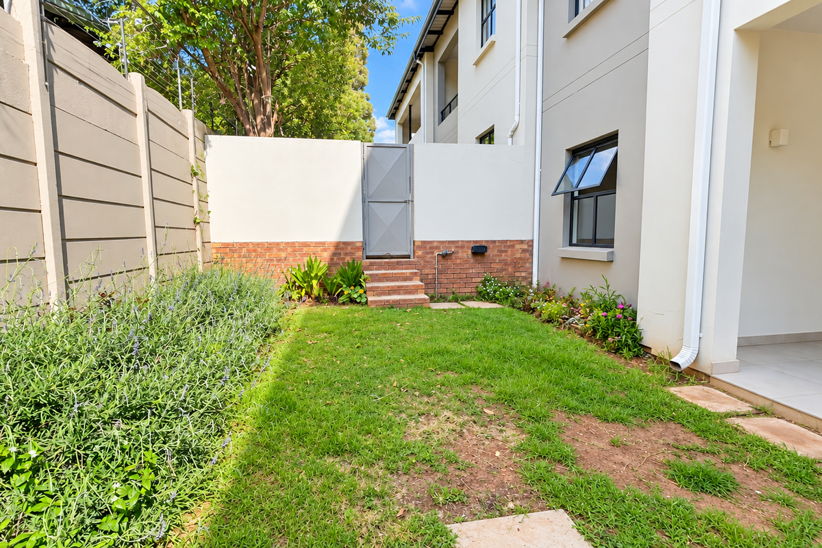 A private rear garden featuring a small lawn, brick-accented boundary wall with a pedestrian gate, and a covered tiled patio area.