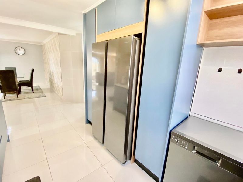 Modern kitchen area featuring sleek blue cabinetry, integrated fridge housing, and light-colored tiled flooring, seamlessly flowing into the adjacent dining space.