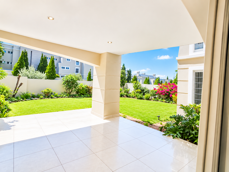 A spacious covered patio with tiled flooring and recessed lighting, overlooking a well-maintained private garden and surrounding townhouse estate.