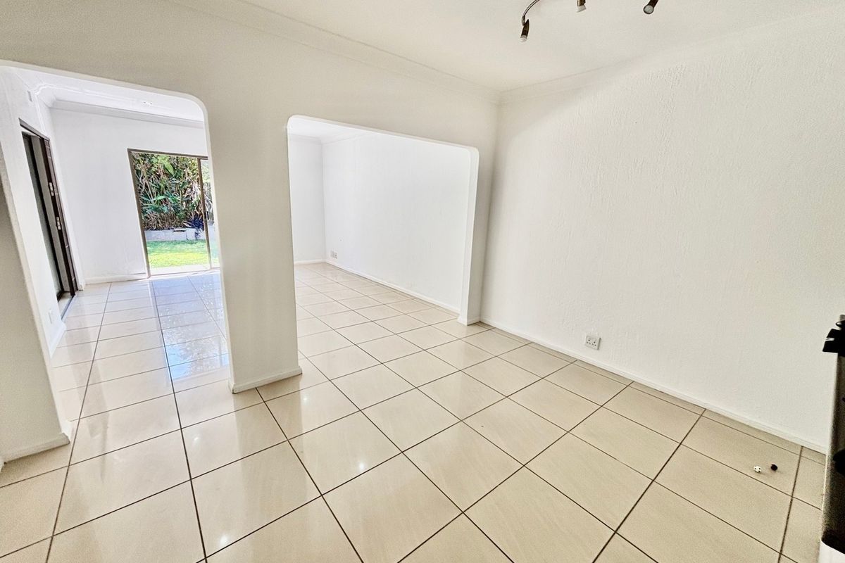 An empty, open-plan living area featuring neutral tiled flooring, white walls, and an archway leading to an adjacent space with garden access.