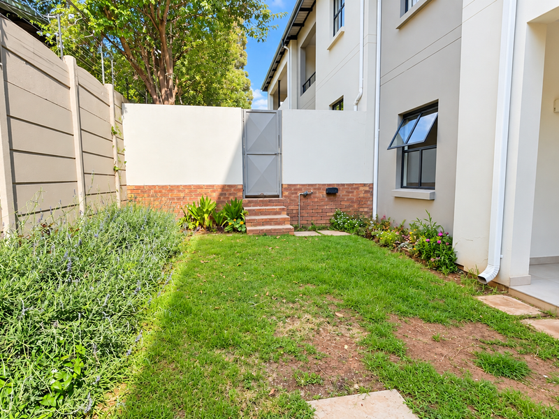 A private rear garden featuring a small lawn, brick-accented boundary wall with a pedestrian gate, and a covered tiled patio area.