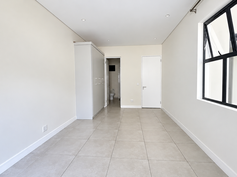 A bright, modern bedroom featuring neutral tiled flooring, built-in wardrobe storage, and a large black-framed window allowing for ample natural light.