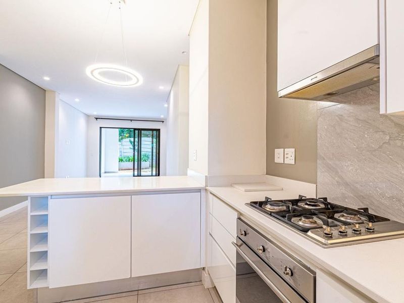 Modern open-plan kitchen featuring sleek white cabinetry, stone countertops, gas hob, and integrated oven, flowing seamlessly into a bright living area with sliding doors.