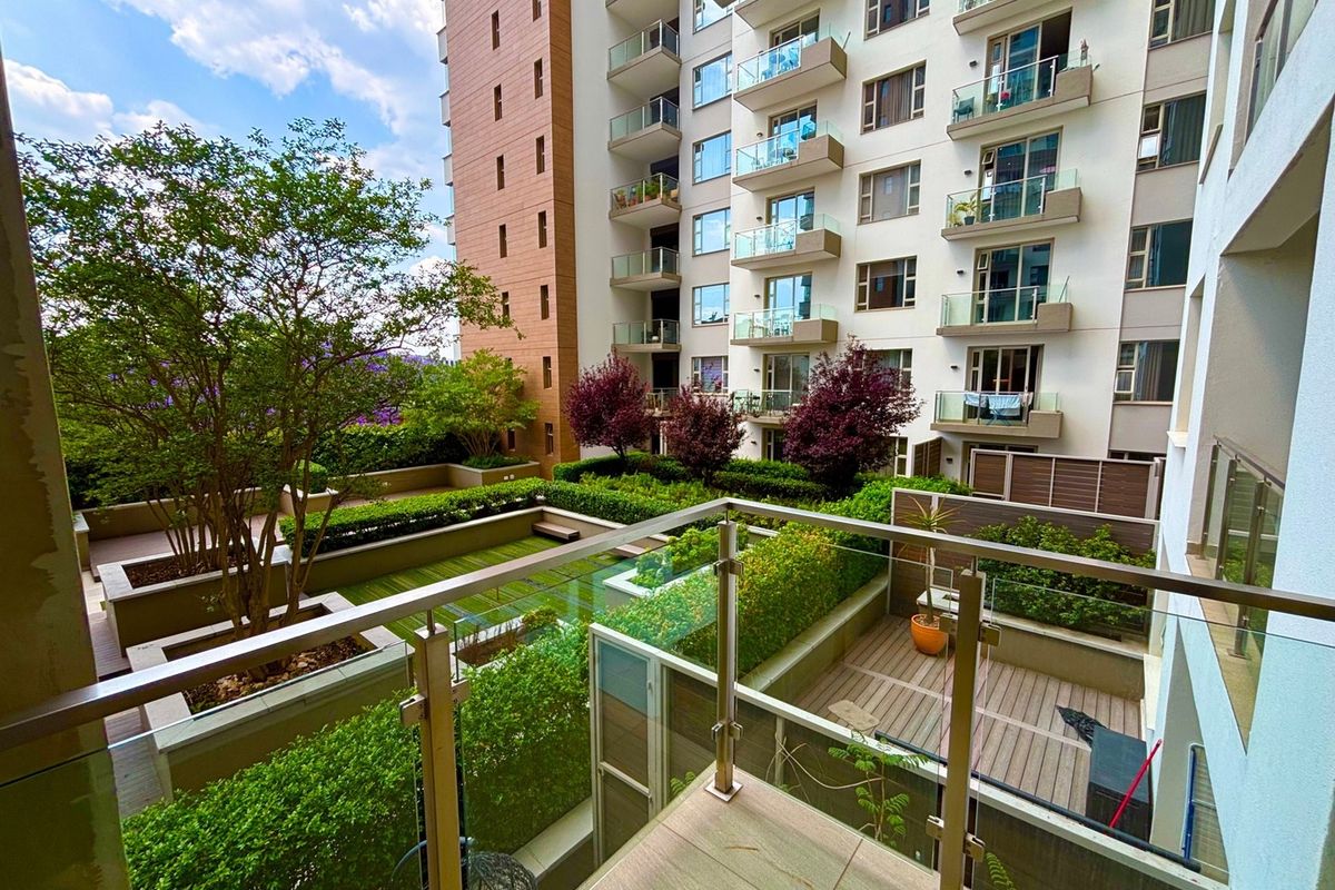 A view from a balcony overlooking a well-maintained communal garden area with timber decking, landscaped hedges, and modern apartment building architecture.