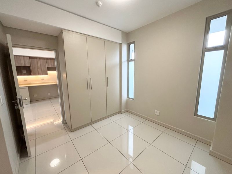 A bright, modern bedroom featuring neutral walls, polished tiled flooring, and a spacious built-in wardrobe, with a view into the adjacent kitchen area.