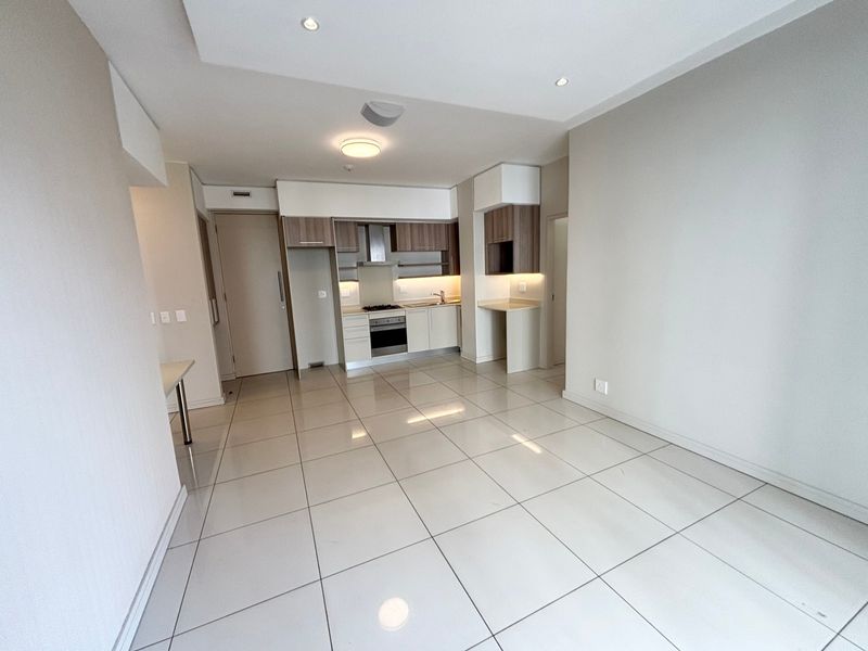 A bright, open-plan living and kitchen area featuring modern cabinetry, integrated appliances, tiled flooring, and recessed lighting in a clean, contemporary apartment setting.