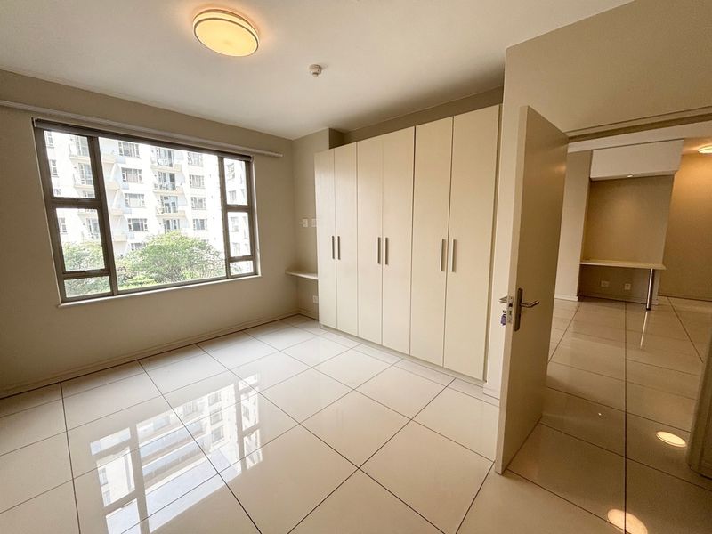 A bright, empty bedroom featuring large windows, polished tiled flooring, and ample built-in cupboard storage space.