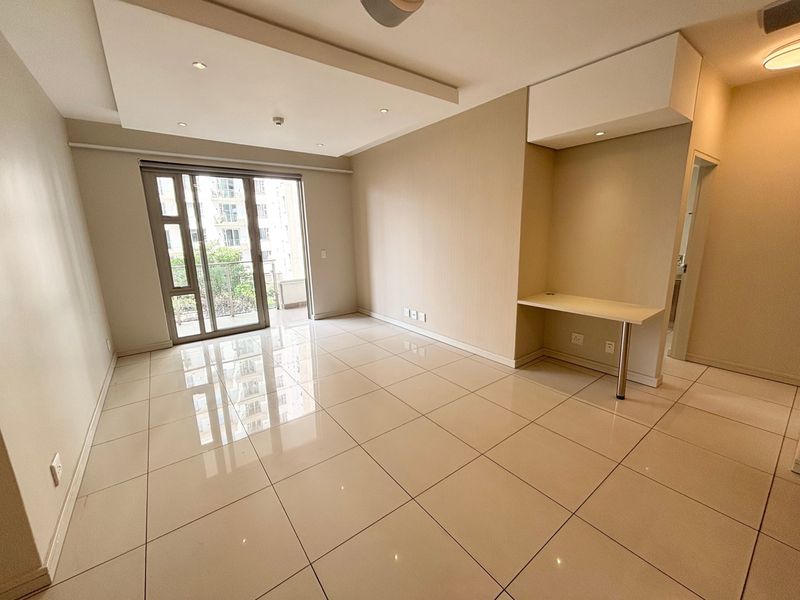 A bright, open-plan living area featuring polished porcelain floor tiles, a modern recessed ceiling with downlights, and a convenient built-in desk nook.