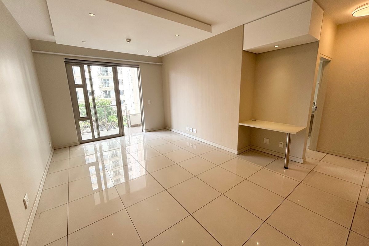 A bright, open-plan living area featuring polished porcelain floor tiles, a modern recessed ceiling with downlights, and a convenient built-in desk nook.