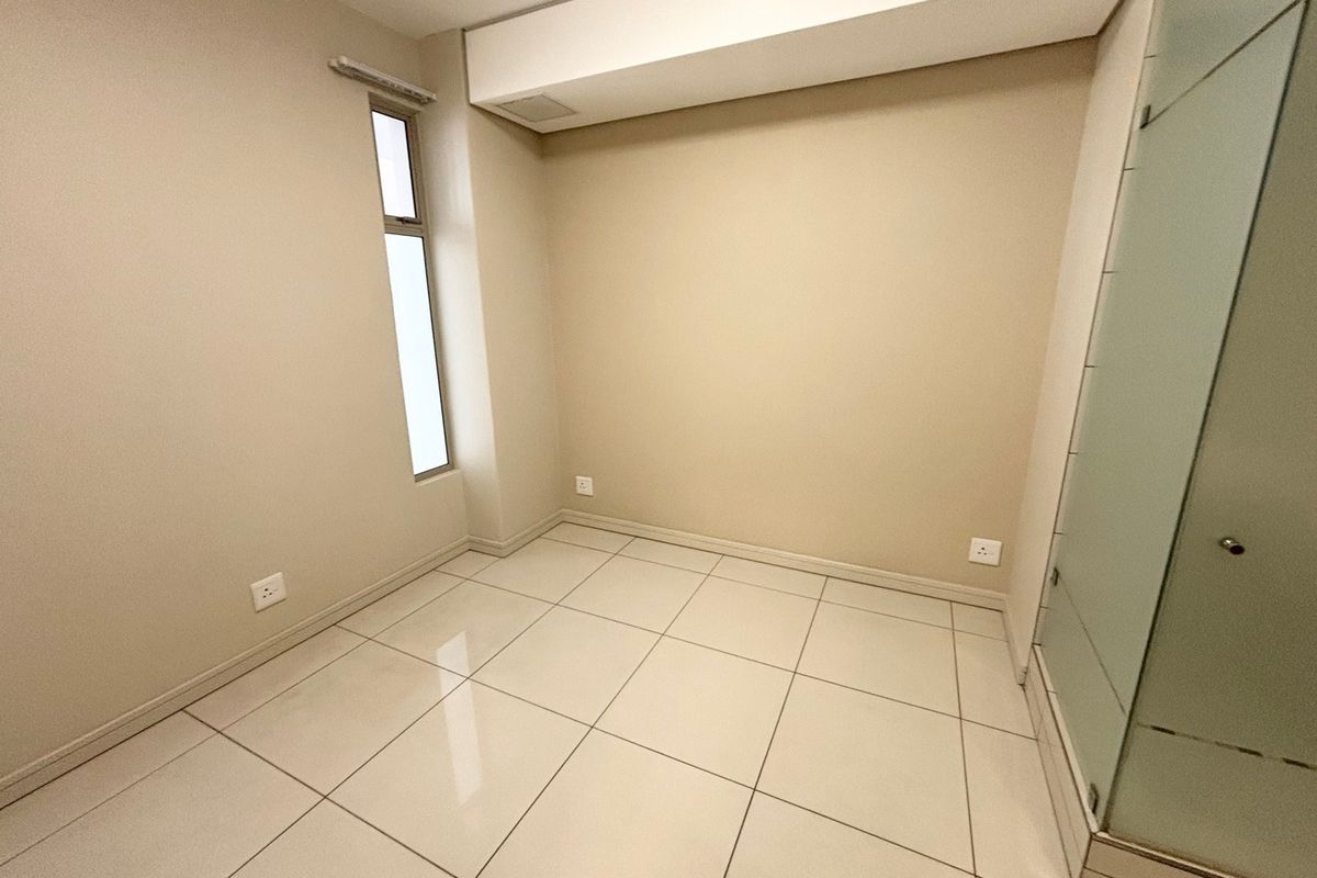 A clean, empty bedroom featuring neutral tiled flooring, a vertical window, and a modern frosted glass door, providing a bright and versatile living space.