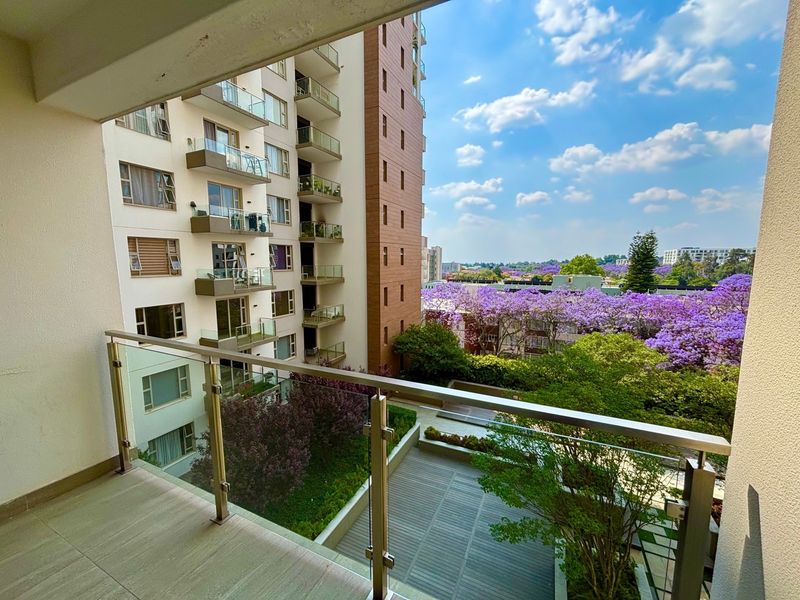 A modern apartment balcony featuring a glass balustrade with stainless steel handrails, offering an elevated view of the surrounding landscaped gardens and neighborhood.