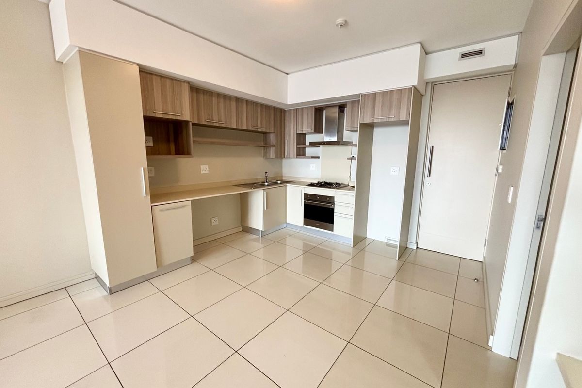 A modern, empty kitchen featuring light wood-tone cabinetry, integrated appliances including an oven and hob, and neutral tiled flooring throughout.