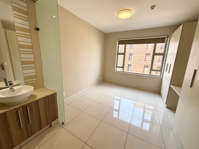 A bright, empty bedroom featuring tiled floors, a built-in vanity with a vessel sink, glass partition, and integrated wardrobe with a desk area.