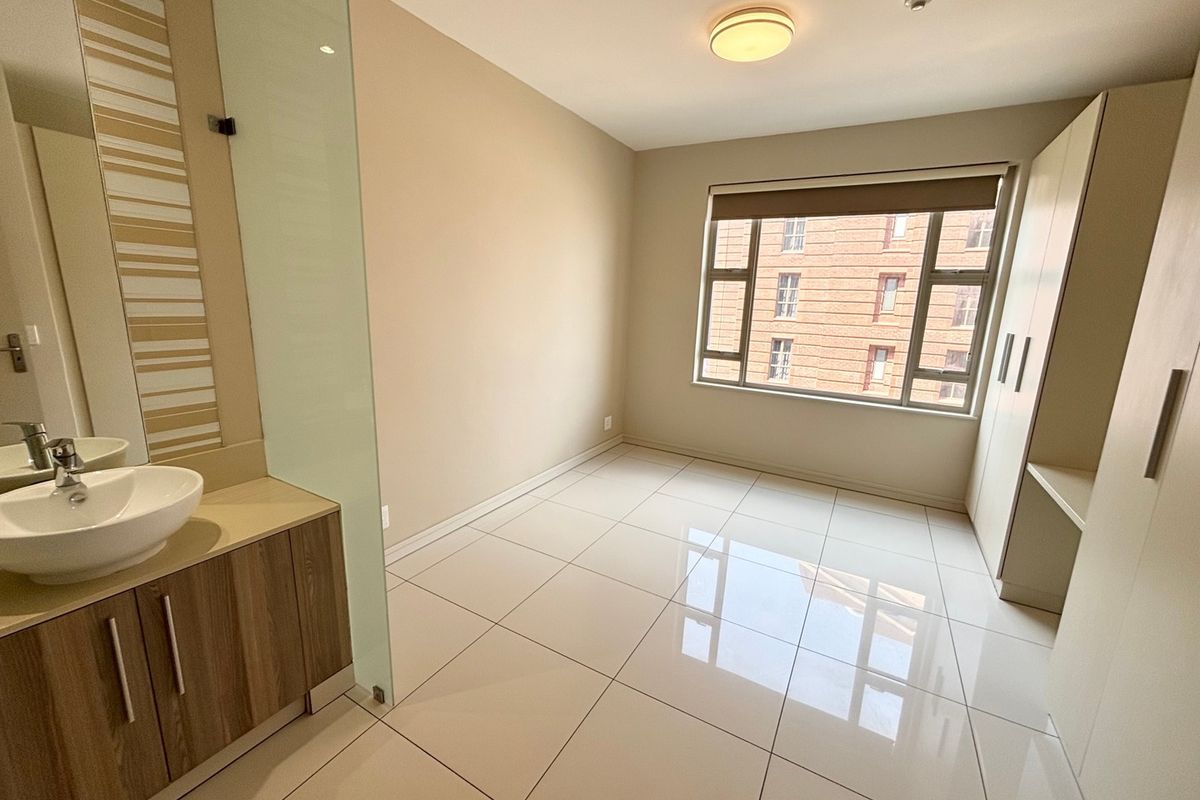 A bright, empty bedroom featuring tiled floors, a built-in vanity with a vessel sink, glass partition, and integrated wardrobe with a desk area.