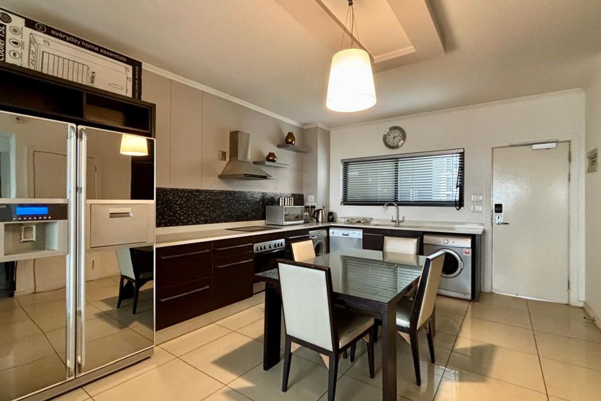Modern open-plan kitchen and dining area featuring dark cabinetry, tiled floors, integrated appliances, and a stylish recessed ceiling with pendant lighting.