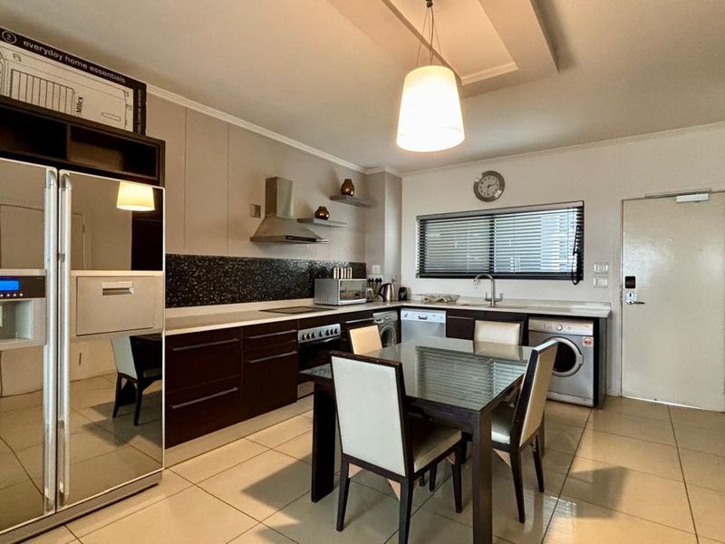 Modern open-plan kitchen and dining area featuring dark cabinetry, tiled floors, integrated appliances, and a stylish recessed ceiling with pendant lighting.