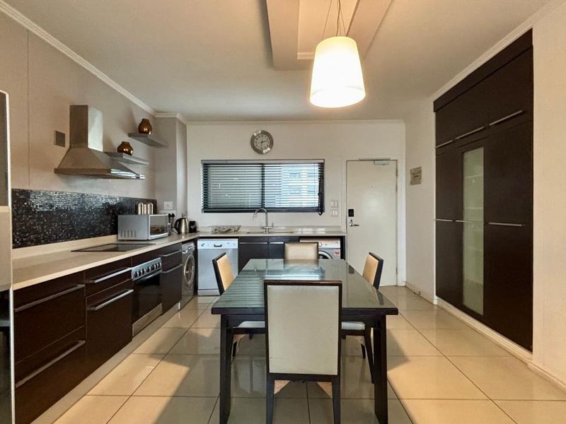 Modern open-plan kitchen and dining area featuring dark cabinetry, mosaic backsplash, tiled flooring, and integrated appliances in a well-maintained apartment setting.