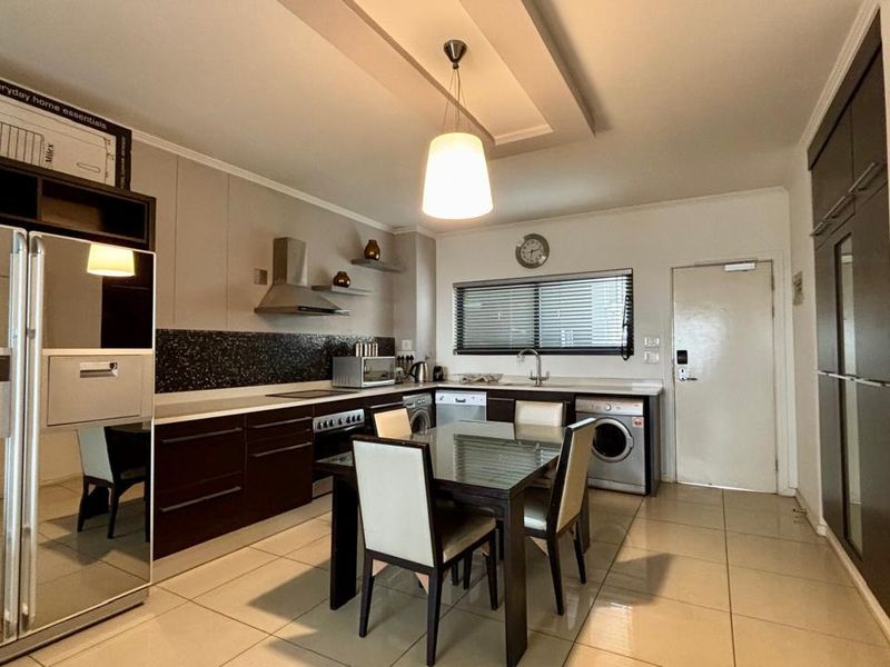 Modern open-plan kitchen and dining area featuring dark cabinetry, integrated appliances, tiled flooring, and a contemporary recessed ceiling design.