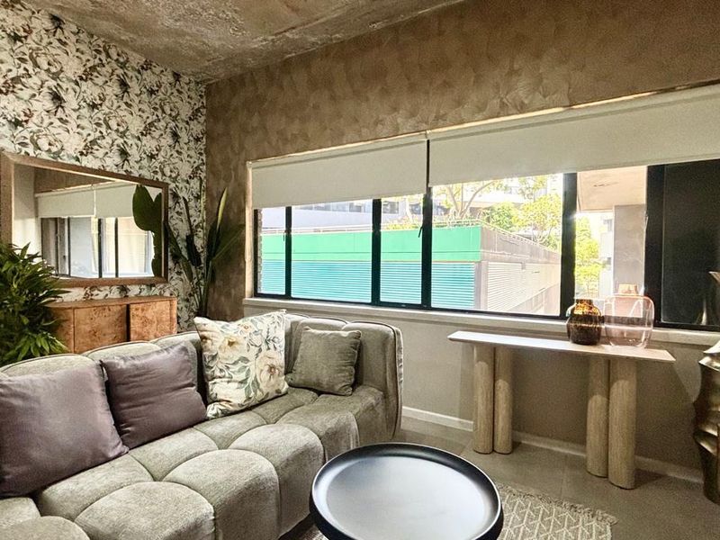 A well-lit living room featuring an exposed concrete ceiling, decorative wallpaper, and large windows with roller blinds, creating a modern and textured aesthetic.